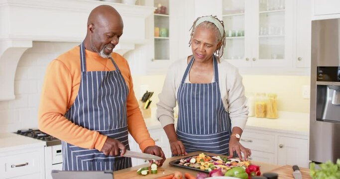 African american senior couple in aprons preparing meal using tablet in kitchen, slow motion