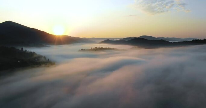 Aerial survey of mountains covered with green forests and spruce with fog or smoke between them in summer weather. The relief surface of the Earth with forests, meadows and small villages, settlements