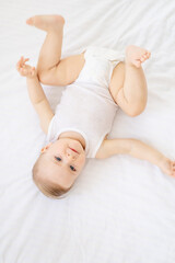 happy little baby girl or boy in a white bodice on the bed in the bedroom looking up, baby with blue eyes
