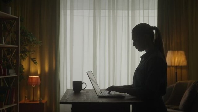 Side view of a dark silhouette of a woman at a table in front of a laptop. A woman works on a laptop with a cup of coffee, typing, thinking. HDR BT2020 HLG Material.