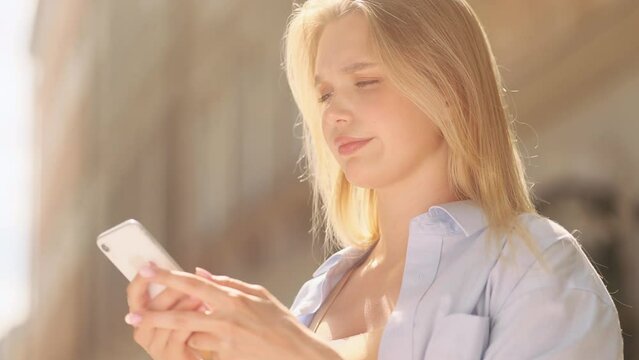 Irritated young blond woman texting at mobile phone at city centre Nervous female typing on smartphone expressing negative emotion outdoors Bad conflict working or personal conversation Fraud