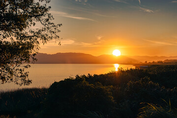 Majestic sunset behind mountains reflected in a serene lake, surrounded by bushes and a solitary...