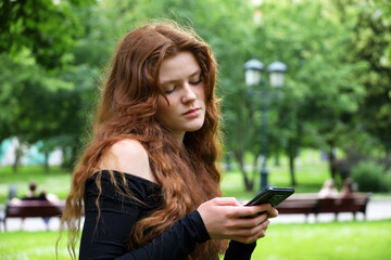 Girl with long red hair and freckles using mobile phone standing in summer park