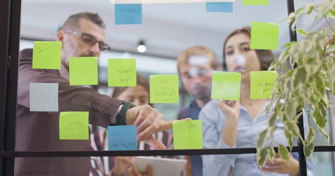 Work colleagues discussing startup project ideas. Team of caucasian business people standing near glass wall with sticky notes. Scrum methodology with colorful stickers. Low angle view focus on window
