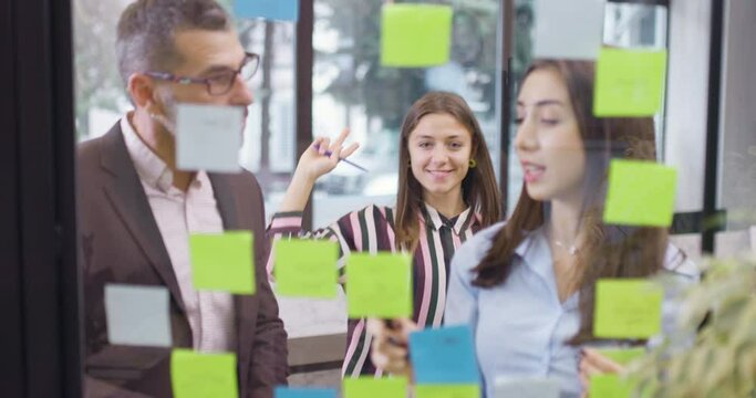 Work colleagues discussing startup project ideas. One happy girl dancing and fooling in the background. Caucasian business team at glass wall with sticky notes. Scrum with colorful stickers.