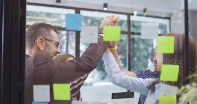 Work colleagues discussing startup project ideas and giving high five. Caucasian business team at glass wall with sticky notes. Scrum with colorful stickers. Vertical movement.