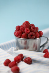 fresh summer sweet and healthy raspberries in a tin mug on a pale blue background and with a striped blue cloth napkin next to it. for postcards, flyers, banners, splash screens, stickers, etc.