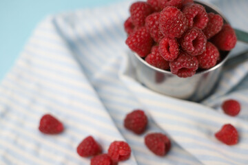 fresh summer sweet and healthy raspberries in a tin mug on a pale blue background and with a striped blue cloth napkin next to it. for postcards, flyers, banners, splash screens, stickers, etc.