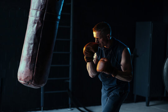 Male boxer in boxing gloves training on punching bag in dark gym.