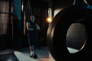 Fototapeta na wymiar Tired boxer resting in dark gym after exercising near punching bag.