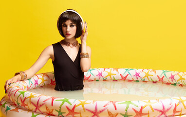 Beautiful young woman in image of Cleopatra sitting in swimming pool and listening to music in headphones against yellow studio background. Concept of antique culture, history, comparison of eras, art