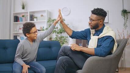 African American psychologist and a happy boy giving each other high five after successful session - obrazy, fototapety, plakaty