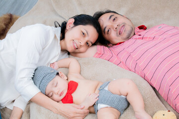 Asian parents lying and looking at camera next to newborn baby boy  deeply asleep in bed. Happy...