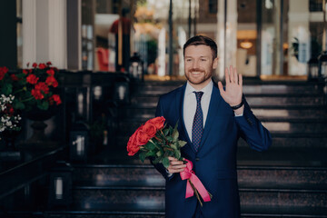 Attractive man in elegant blue suit waits on street with rose bouquet, greeting date in romantic...