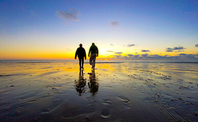 Naklejka premium An evocative photograph capturing the silhouette of two teenage girls walking on a beach, set against a dramatic and colorful sky. The vibrancy of the sky is beautifully reflected on the water and