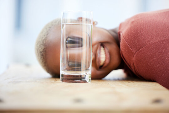 Woman, funny face and water glass with big eyes, laughing and comic joke on table in apartment. African girl, crazy young student and comedy for meme with happy smile, home and playing game on desk