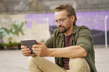 Mid-aged man engaged in studying from a tablet PC while sitting outdoors. With a focused expression, he utilizes the digital device to access study materials and engage in self-improvement.