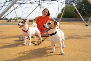 Summer walk with two dog. Teen girl in orange clothes sitting playground with her small cute white Jack Russell terriers pets with double leash. Summer in big cityю girl in the background out of focus