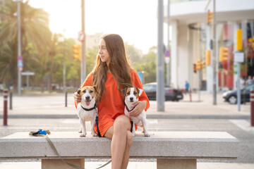 Girl with two dog friends. Summer time vibe in big city. Smiling happy teenage girl profile in orange clothes looking side hugging cute pets Jack Russells sitting on the bench. 