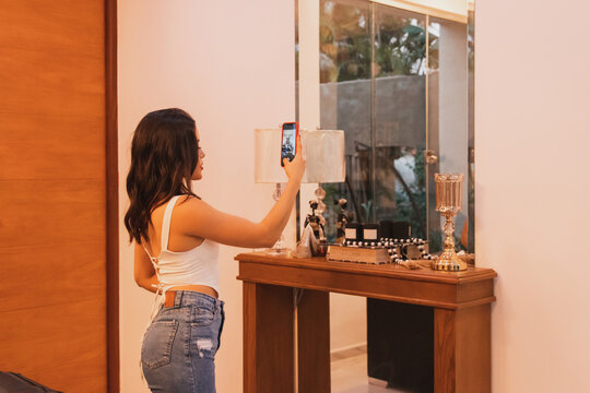 Beautiful young girl taking pictures of herself with a smartphone in the mirror in her living room.