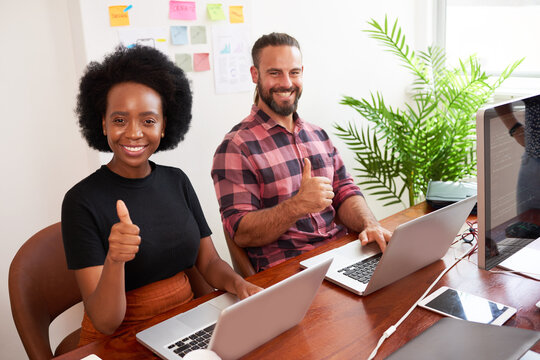 Two developers sitting at shared desk give thumbs up, coding in office on laptop