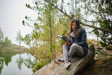 latina woman reading a book in a forest with a lagoon in the mountains of bolivia