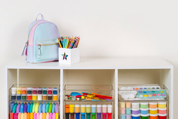 White shelving with kids backpack and various material for creativity and art activity. Stationery and supplies for drawing and craft. Organizing and storage in childrens room.