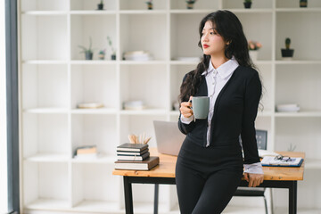 Happy young Asian businesswoman leader hand holding coffee cup, standing in an office. Professional executive manager or saleswoman using corporate technology.