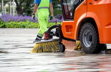 The sweeping machine brushes the sidewalk. A worker with a broom is walking in front of the car.