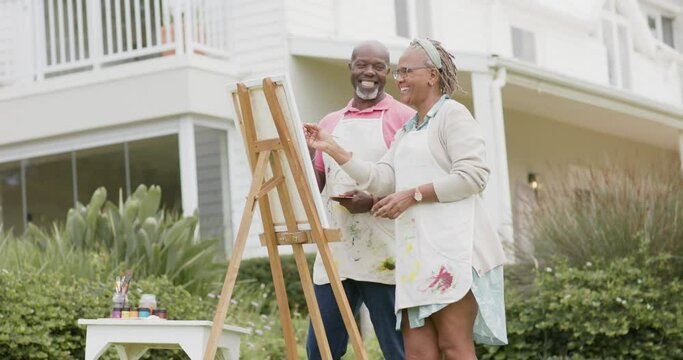 Happy senior african american couple painting on wooden easel in garden, slow motion