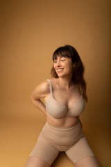 Studio portrait of a caucasian woman in her 30s wearing nude colour shapewear and a bra. The studio background is beige.