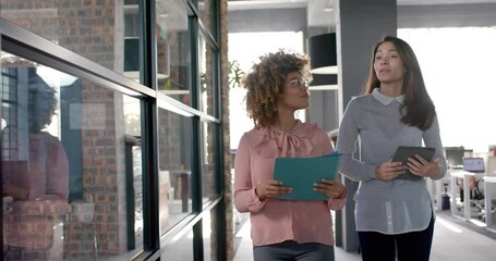 Happy biracial female colleagues walking through office talking, with tablet and folder, slow motion