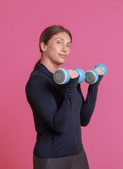 Workout concept. Young athletic woman in sportswear trains biceps muscles with dumbbells in her hands on pink background. Bodybuilding and fitness. Healthy lifestyle