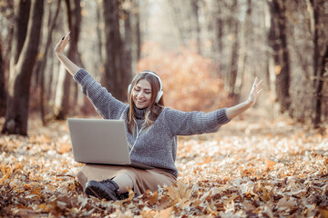 Young woman in headphones with a laptop sits on fallen leaves in autumn forest and has fun listening to music