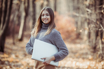 Portrait of a cute Caucasian woman in a warm sweater with a laptop in her hands in the autumn forest. Remote work or freelancing concept