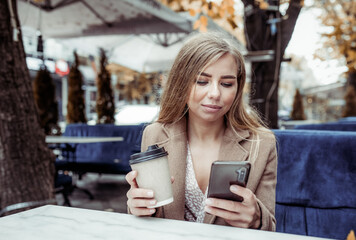 Young beautiful woman in a coat drinking coffee and using smartphone in outdoor cafe