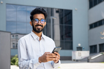 Obraz na płótnie Canvas Portrait of young indian programmer businessman, man in glasses holding phone, looking at camera and smiling, happy with achievement results freelancer.