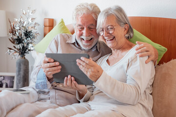 Cheerful senior family couple together in bed, two elderly people laughing relaxing at home using digital tablet technology, leisure time