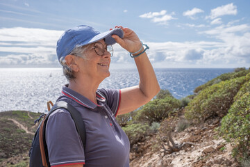 Active senior woman in blue hat walking on trail outdoors on country excursion. Scenic seascape on background. Happy caucasian woman enjoying travel or retirement leading healthy lifestyle.