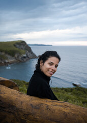 Young hispanic woman smiles sitting on a bench over the beach in cold weather