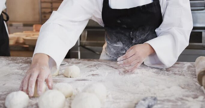 Midsection of asian female baker working in bakery kitchen, forming rolls from dough, slow motion