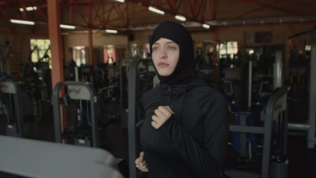 Young muslim female athlete in a hijab running on a treadmill in an indoor gym. Sportswoman in black sportswear doing exercises during cardio workout
