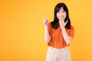 Cheerful Asian woman 30s wearing orange shirt pointing finger to free copy space while gently closing her mouth with hand against yellow background, capturing attention and adding intrigue concept.