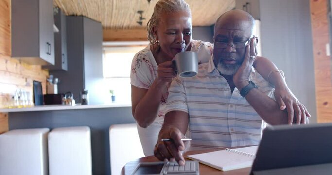Senior african american couple embracing and doing paperwork using tablet at home, slow motion