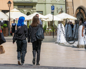 Two young female friends with fashionable hairstyles hold hands and walk along the street of the old city on a sunny day, a girl with blue hair, lesbians in black suits in love. Street style fashion