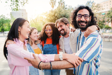 Group of joyful young adult friends embracing each others on a social gathering outdoors, having fun and enjoying standing in a circle. Strong friendship multiracial people hugging showing team unity