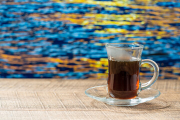 Glass mug of hot black tea on wooden table with sea water during sunset on summer day in tropical beach cafe, closeup, copy space