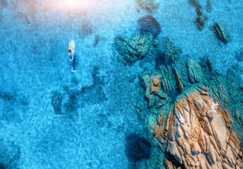 Aerial view of man on sup board in blue sea, rocks at sunrise in summer. Man on floating canoe in transparent azure water. Kayak. Sardinia island, Italy. Tropical seascape. Active travel. Top view