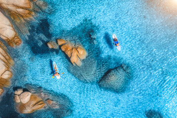 Aerial view of sup boards, blue sea, rocks at sunrise in summer. People on floating canoe in transparent azure water. Kayak. Sardinia island, Italy. Tropical seascape. Active travel. Top drone view - obrazy, fototapety, plakaty