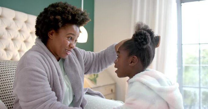 Happy african american mother combing daughter hair sitting on bed in bedroom, slow motion